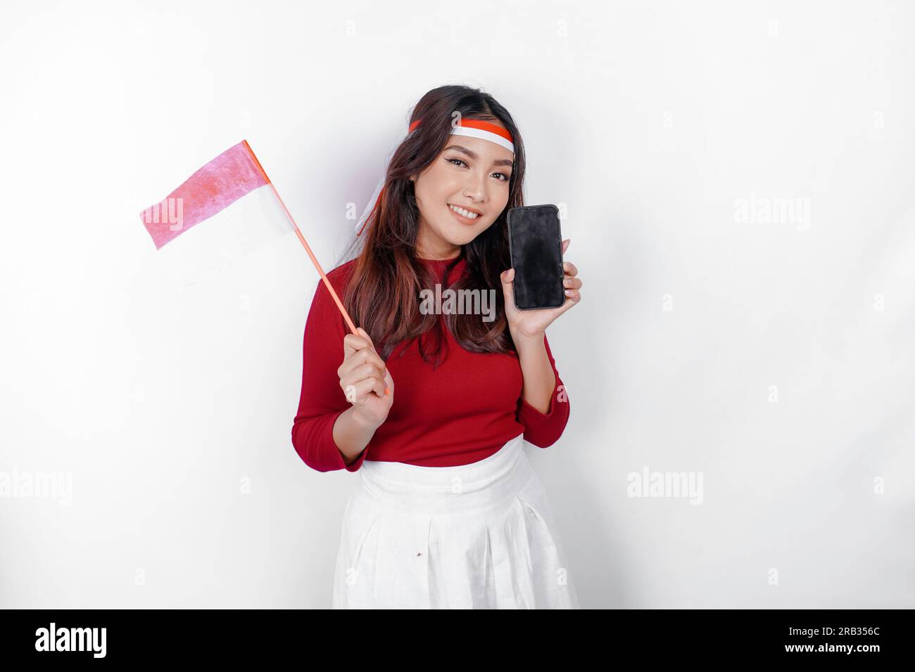 Una felice donna asiatica che indossa un cerchietto, regge il telefono e la bandiera indonesiana, isolata da uno sfondo bianco. Il giorno dell'indipendenza dell'Indonesia Foto Stock