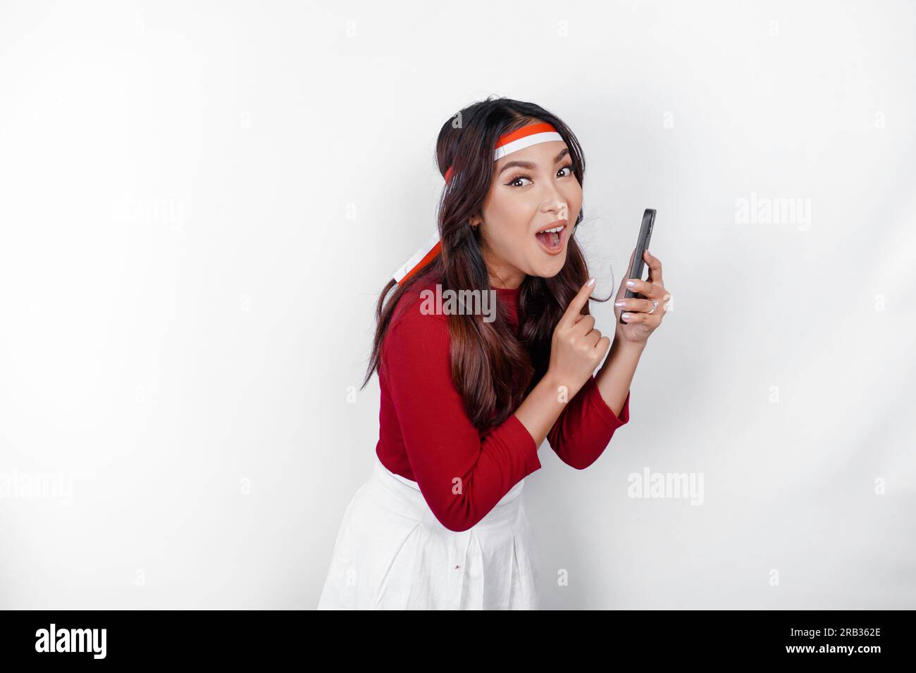 Un ritratto di una donna asiatica scioccata che indossa un cerchietto, che tiene il telefono mentre la bocca è spalancata, isolata da uno sfondo bianco. L'independe dell'Indonesia Foto Stock