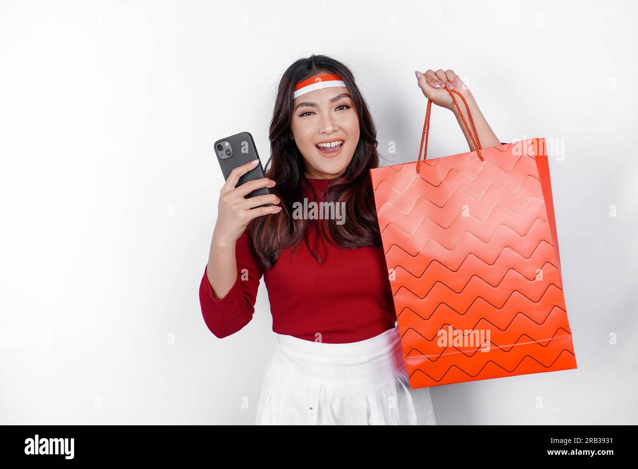 Un'attraente donna asiatica in piedi entusiasta di tenere in mano una borsa per la spesa online e il suo smartphone, lo studio ha girato isolato su sfondo bianco. L'Indonesia e' dentro Foto Stock