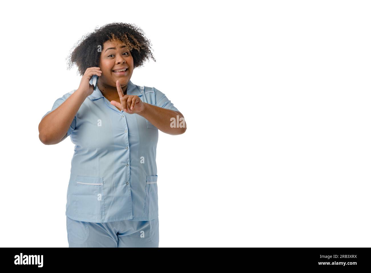 La giovane medico afro-latina in uniforme blu è in piedi sorridente a parlare al telefono con un dito sollevato con un'espressione interrogativa su di lei Foto Stock