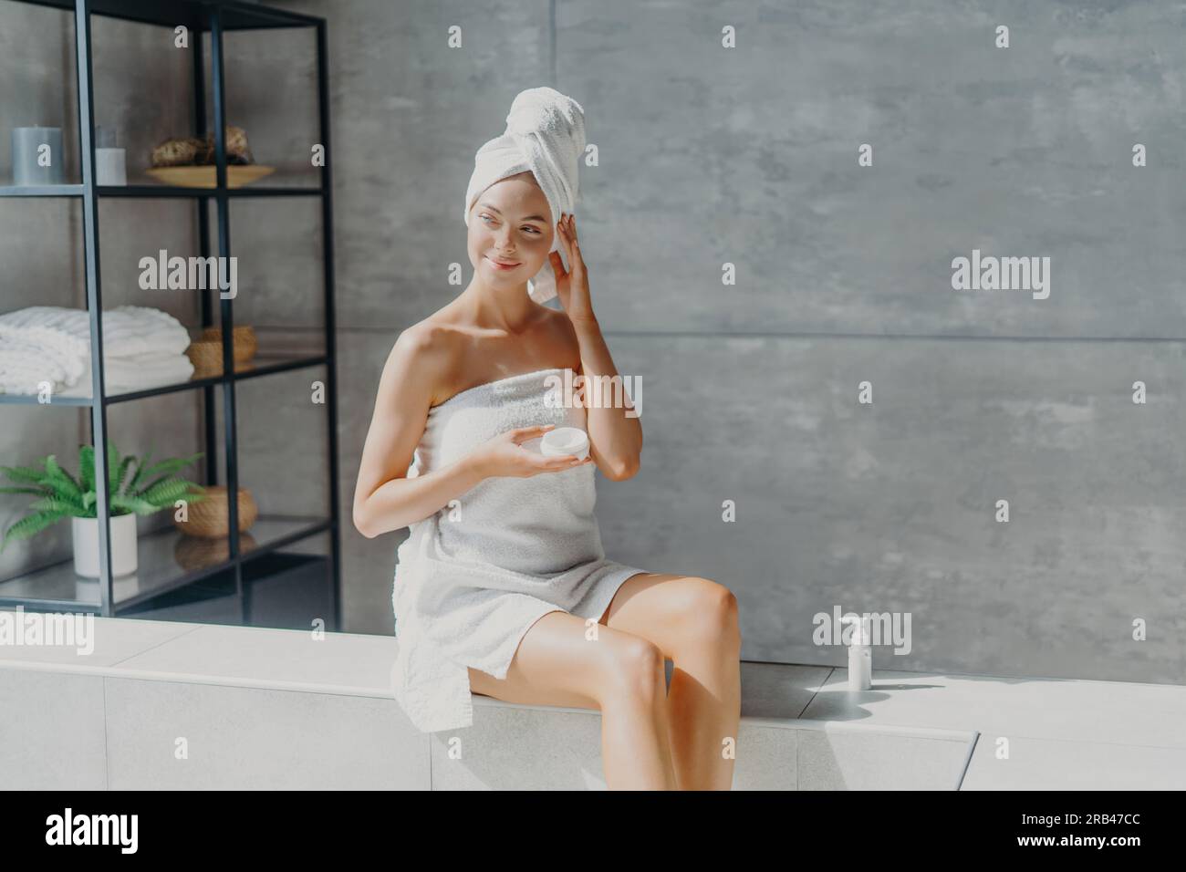 Una donna europea da sogno applica una crema per il corpo, avvolta in un asciugamano. Concetto di salute, bellezza. Viziatevi nel bagno. Foto Stock