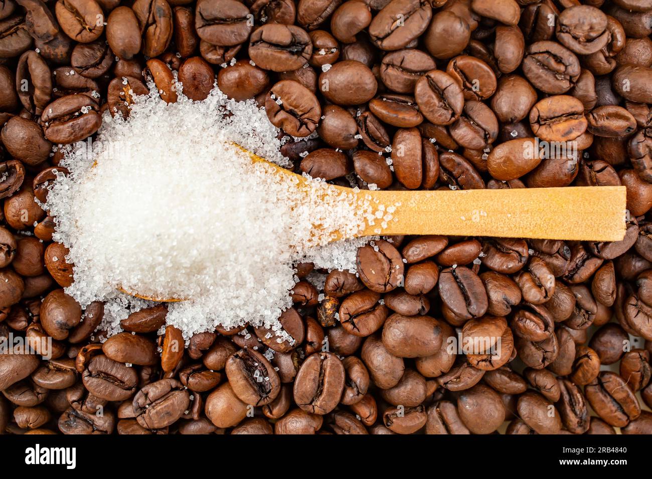 Cucchiaio da tè in legno pieno di cristalli di zucchero, sullo sfondo dei chicchi di caffè, fuoco morbido da vicino Foto Stock