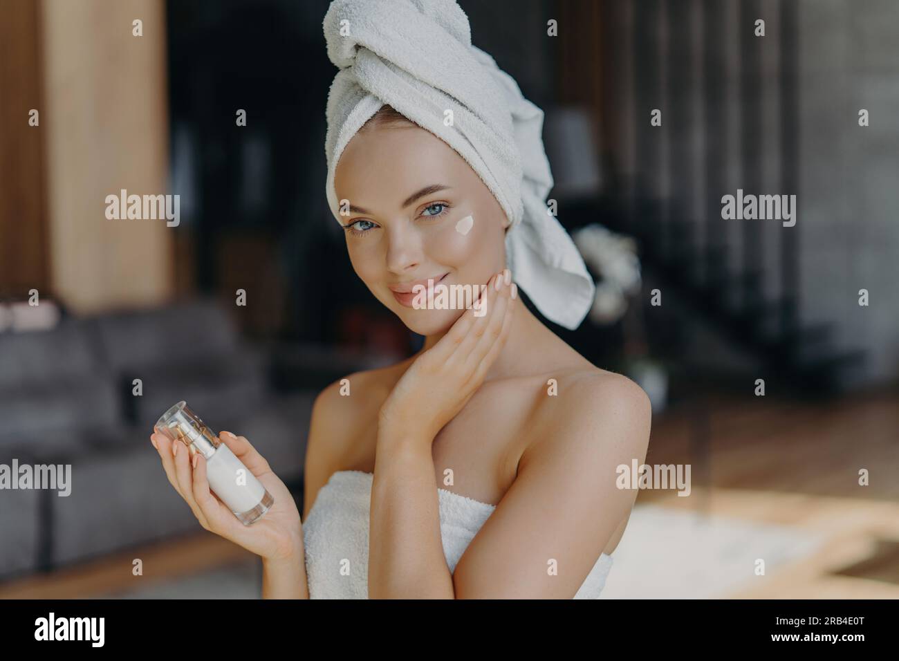 La donna europea applica la crema viso, idrata la pelle. Trucco minimo, asciugamano sulla testa. Interni domestici, trattamento viso. Procedura anti-invecchiamento. Foto Stock
