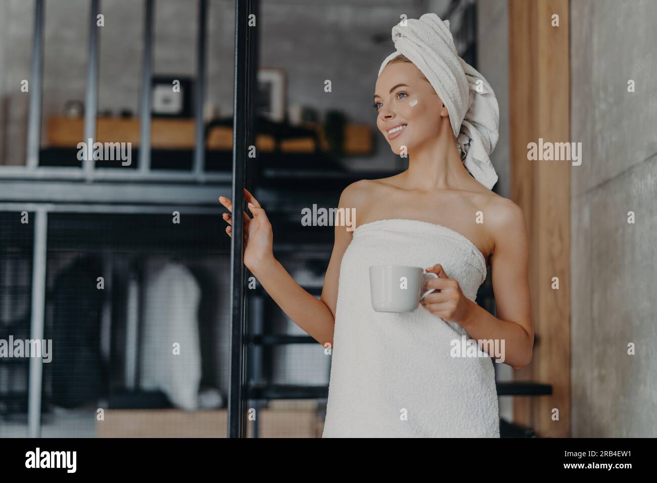 Una donna premurosa sorride, applica la crema, avvolto in un asciugamano, beve tè, si rilassa sulle scale interne dopo il bagno Foto Stock
