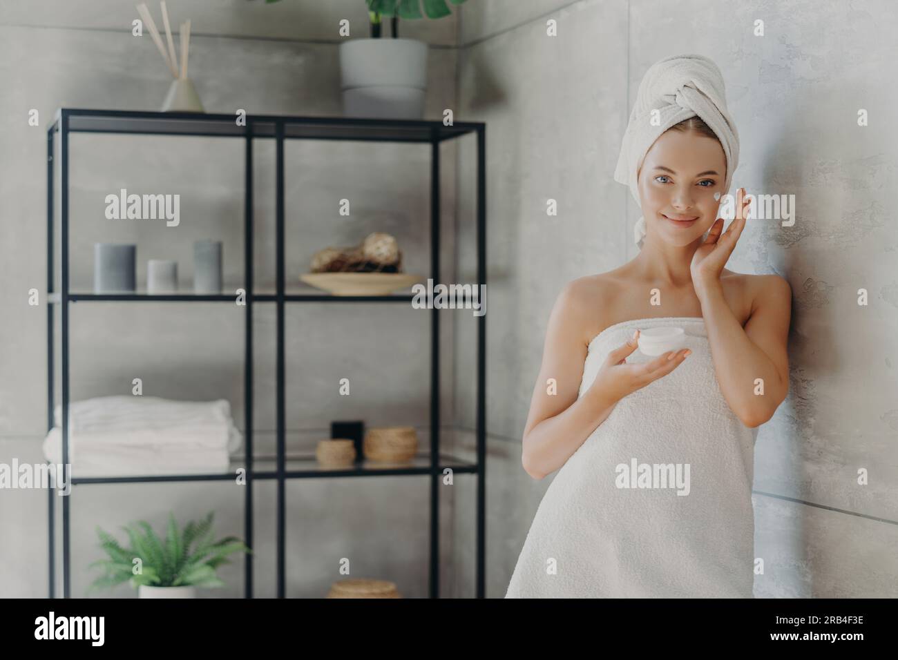 Modello sano utilizza la crema, tiene il vaso cosmetico, avvolto in asciugamano, vicino alla parete accogliente del bagno. Cura della pelle Foto Stock