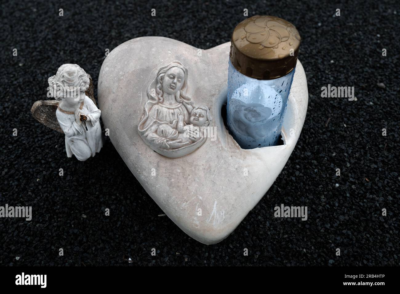 Una pietra a forma di cuore con l'immagine della Beata Vergine Maria e del bambino Gesù, fiancheggiata da due piccole statue di angeli su sfondo di sabbia nera Foto Stock