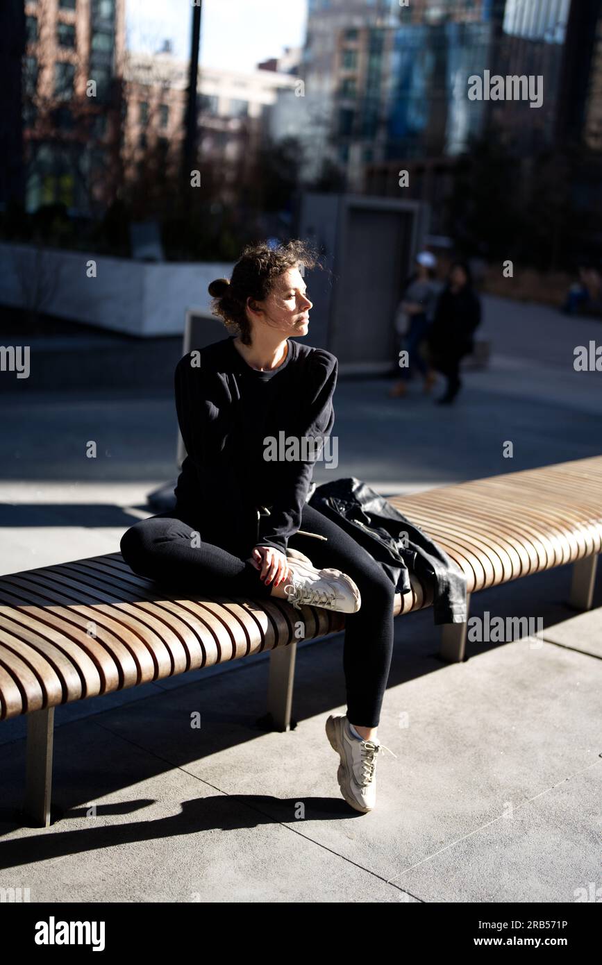 Jogger femminile illuminata dal sole che riposa su una panchina in un ambiente urbano Foto Stock