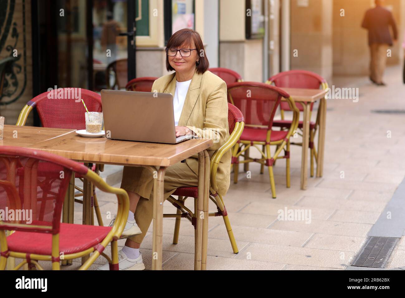Donna d'affari matura ed elegante con occhiali da vista lavora sul computer portatile mentre si siede con un caffè all'aperto in un caffè europeo. Concetto di lavoro remoto da pub Foto Stock