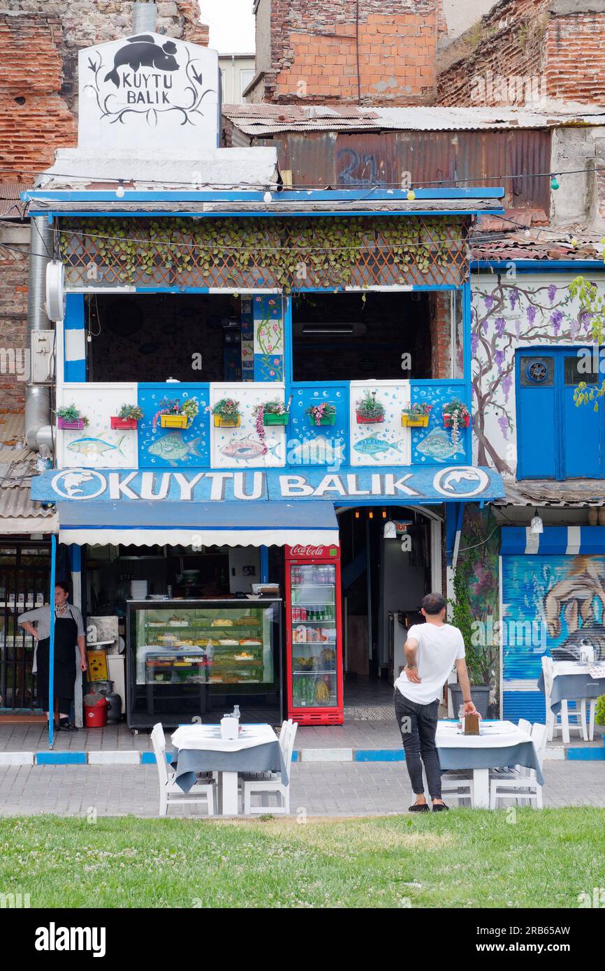L'uomo si arca alla schiena con mal di schiena all'esterno di un caffè blu e bianco con vasi di piante nel quartiere Karakoy, Istanbul, Turchia Foto Stock