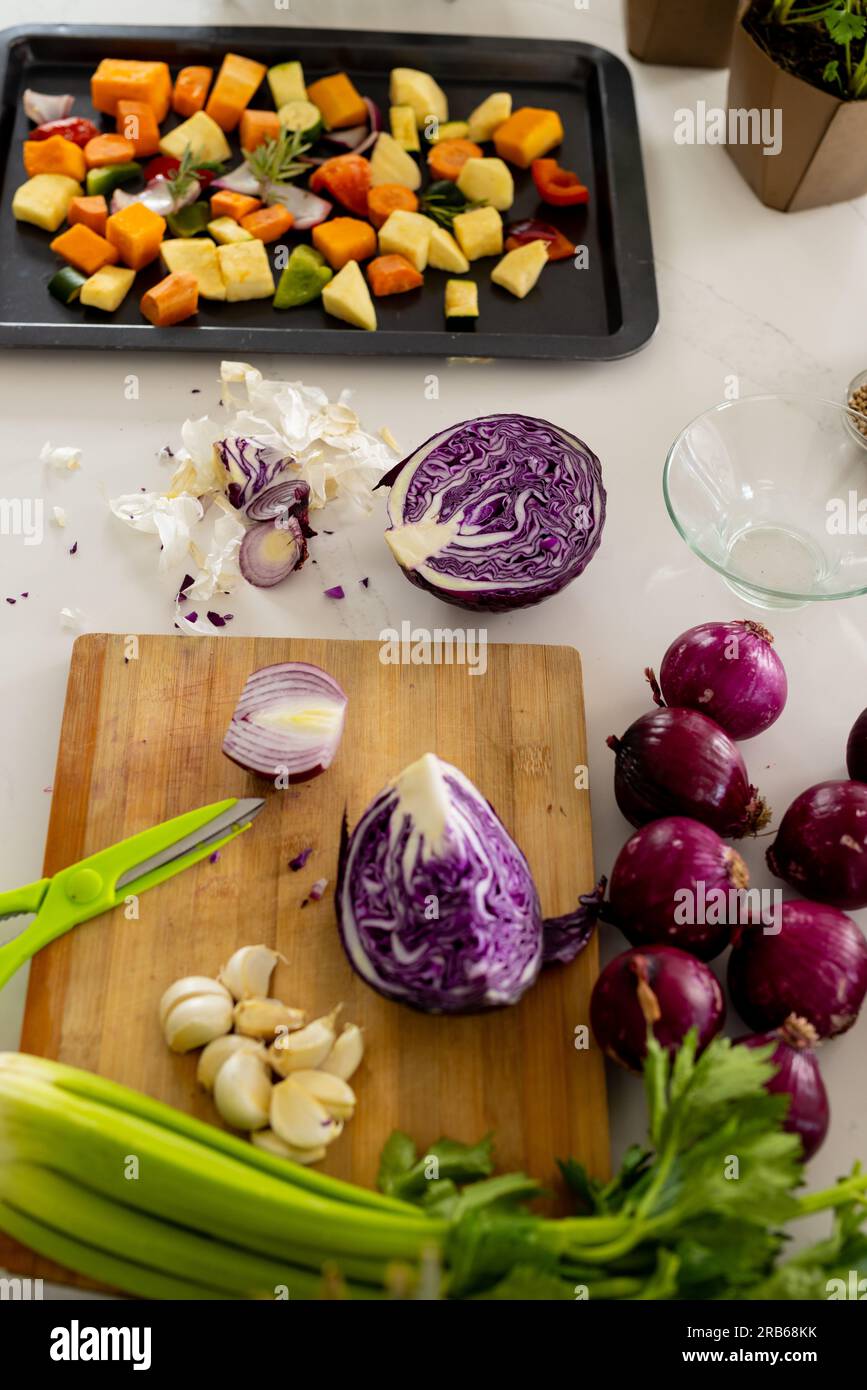 Verdure su tagliere e verdure tritate in vassoio da forno sul piano di lavoro nella cucina soleggiata. Cucina, cibo, ingredienti, vita sana e domesti Foto Stock