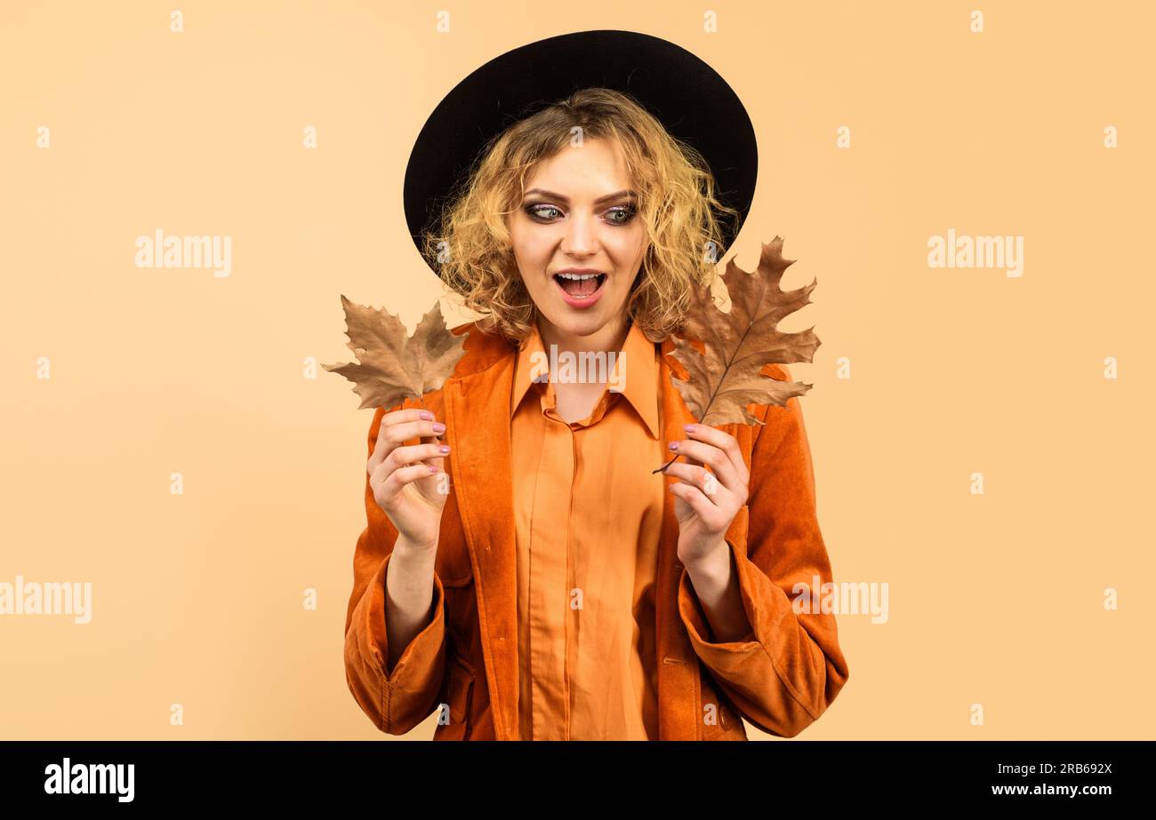 Sorpresa ragazza con cappello nero con foglie autunnali. Donna alla moda con camicia e cappotto arancione con fogliame autunnale. Saldi autunnali. Sconto. Abbigliamento autunnale Foto Stock