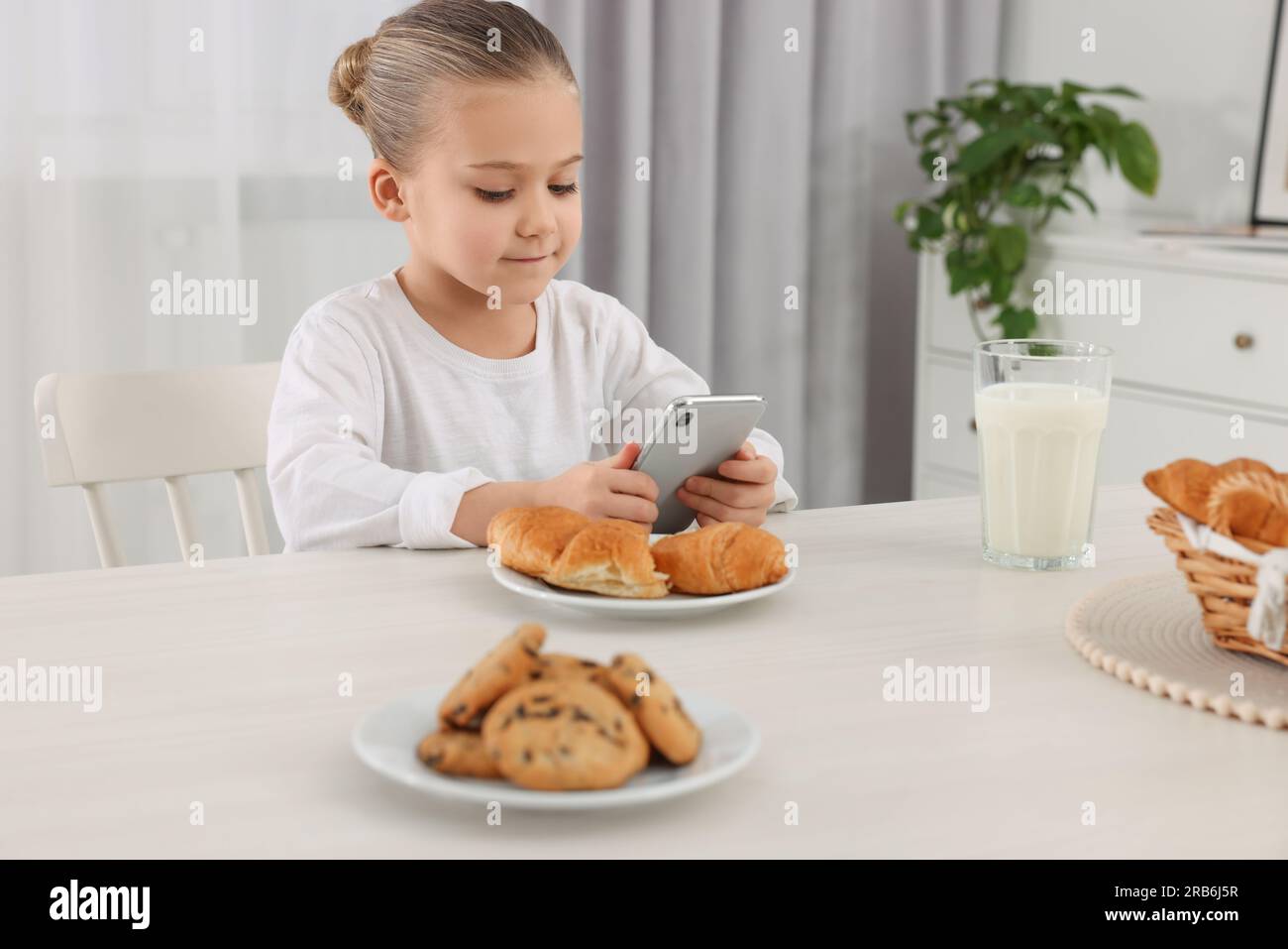 Bambina che usa lo smartphone mentre fa colazione al tavolo all'interno. Dipendenza da Internet Foto Stock