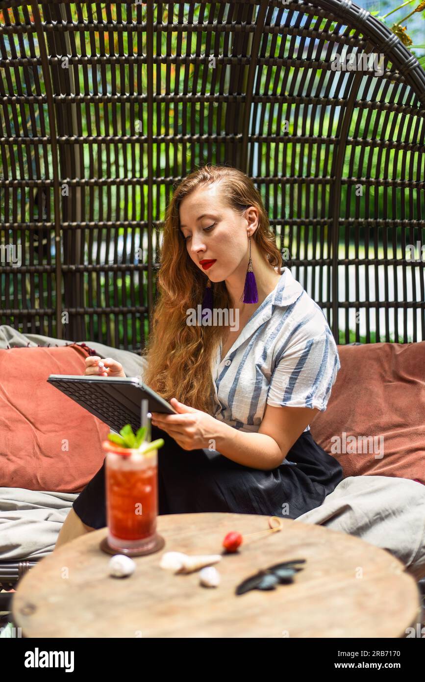 Bella artista e illustratrice bionda che pensa a un concetto del suo disegno, seduta accanto al tavolo con un tablet elettronico e uno stilo Foto Stock