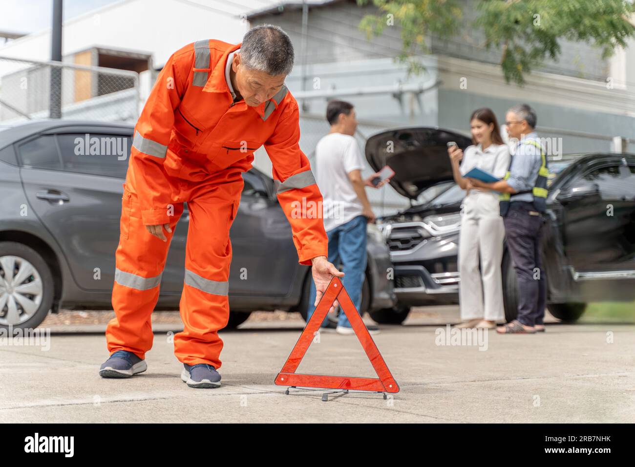 Un agente di assicurazione auto in tuta arancione che posiziona un triangolo segnaletico sulla strada vicino all'incidente mentre un altro agente di assicurazione parla con Th Foto Stock