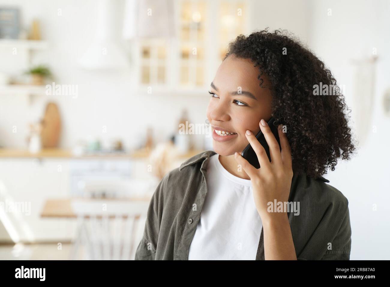 La ragazza amichevole con i capelli afro parla al telefono a casa. L'adolescente sorridente si diverte a conversare piacevolmente al chiuso. Foto Stock