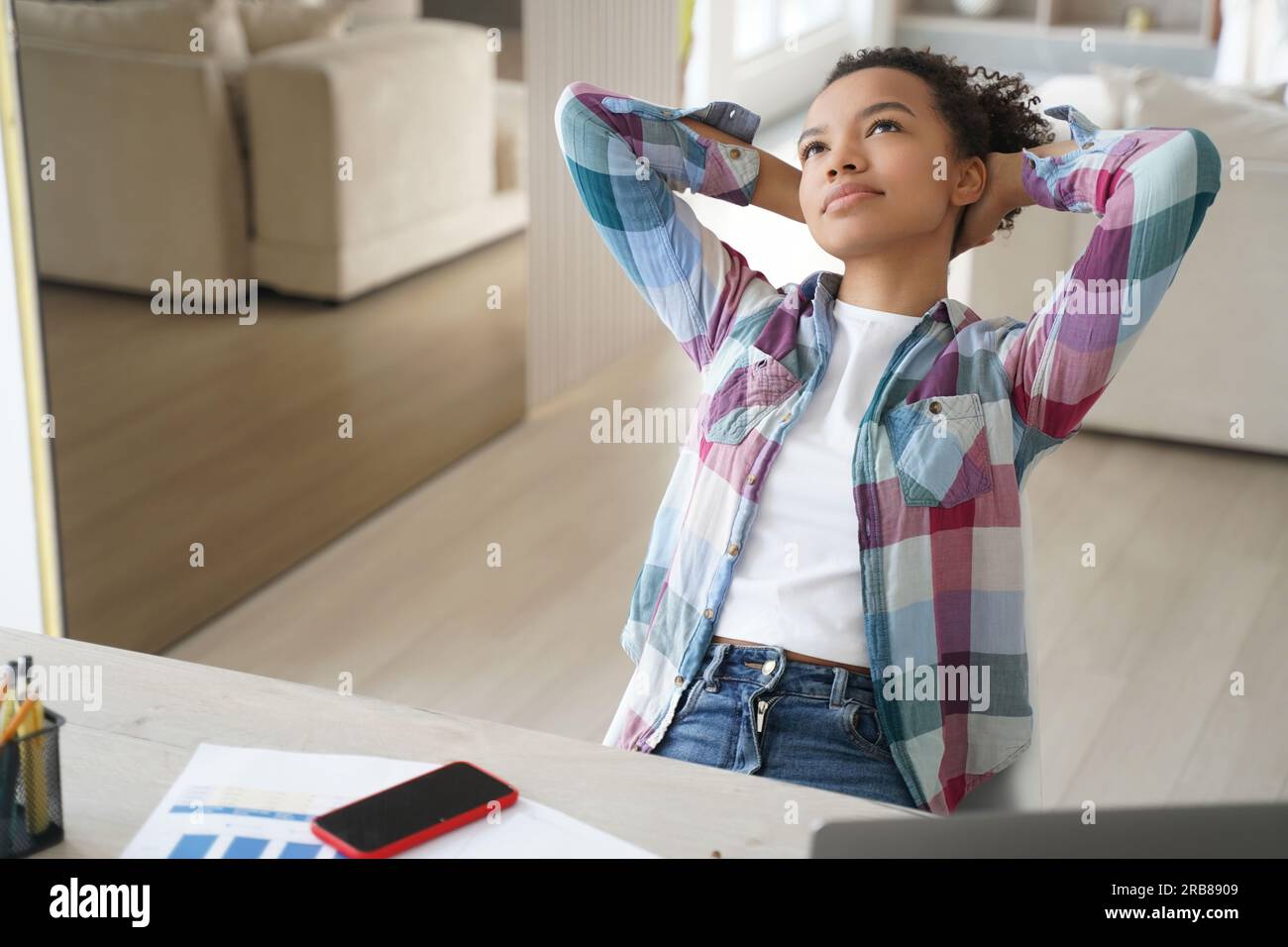 Una studentessa adolescente birazziale sognante si prende una pausa dallo studio a casa, appoggiandosi alla schiena con le mani dietro la testa, perduta nei pensieri. Foto Stock