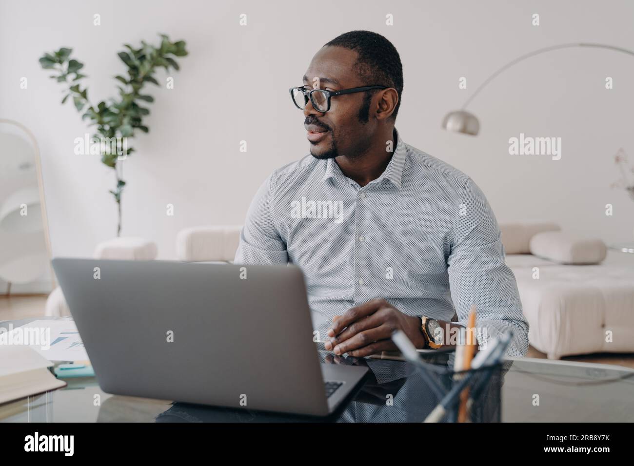 Uomo d'affari afroamericano pensivo che indossa occhiali che lavora su un computer portatile online, pensa a un progetto aziendale, guarda da parte, seduto alla scrivania dell'ufficio. Foto Stock