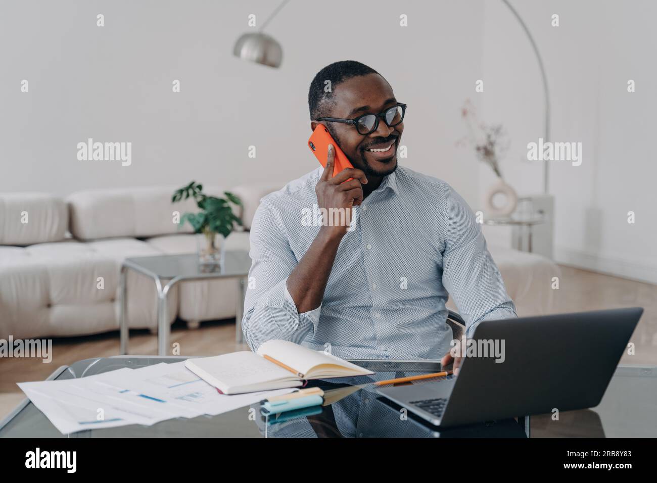 Un uomo d'affari afro-americano felice fa una chiamata d'affari, gode di una conversazione mobile, lavora al computer portatile sul posto di lavoro. Sorridente manager nero discu Foto Stock