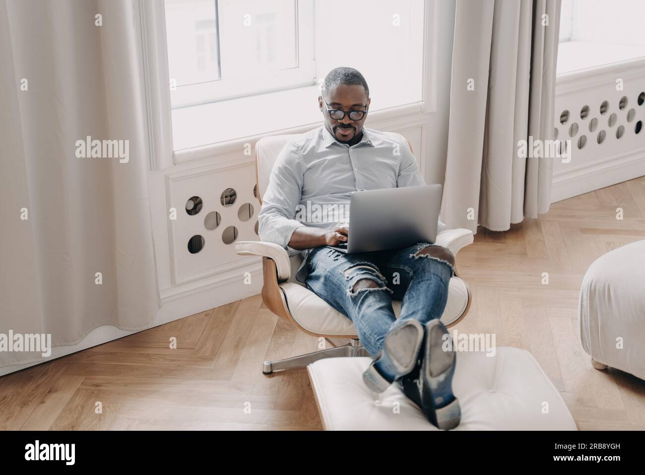 Un uomo d'affari afroamericano concentrato lavora online al computer portatile, seduto in poltrona al chiuso. Moderno dipendente manager nero in occhiali Answers e-mail, Foto Stock