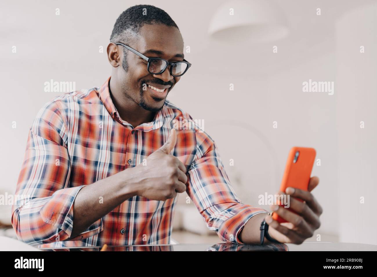 Un uomo afro-americano che mostra il gesto della mano con il pollice in alto, tiene lo smartphone, chatta con una videochiamata. Un ragazzo nero sorridente in occhiali che guarda il telefono Foto Stock