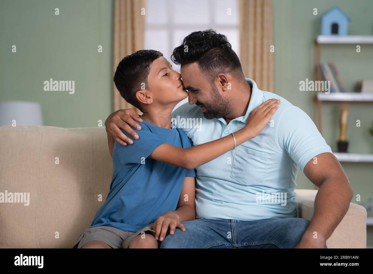 Felice figlio indiano che baciava la fronte del padre mentre sedeva sul divano a casa - concetto di affetto, amore per la famiglia e momento tenero. Foto Stock