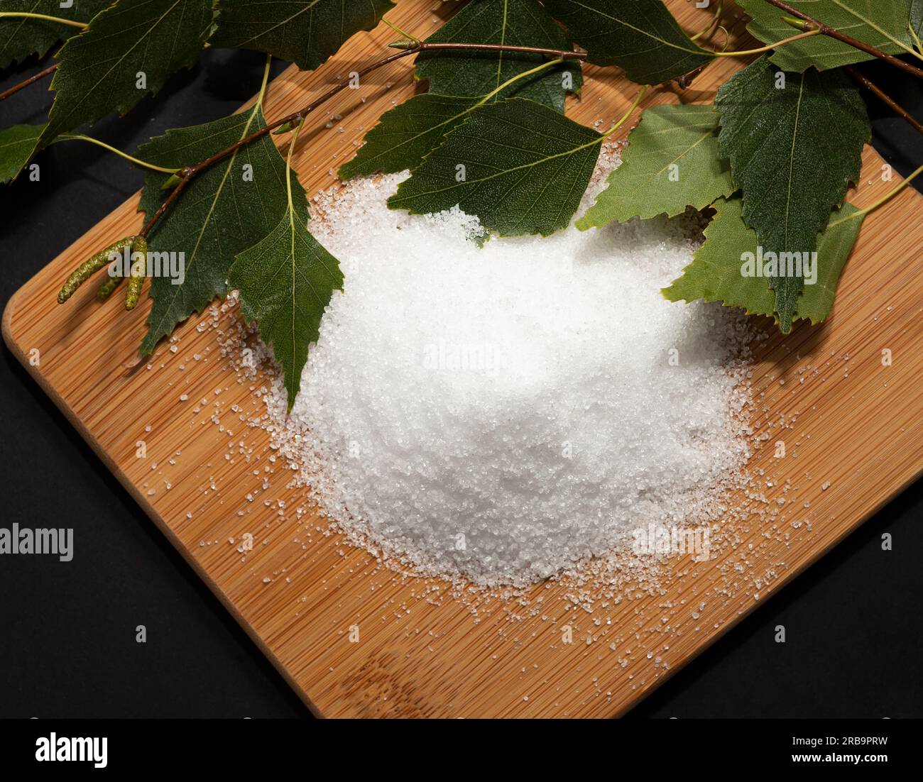 Utile dolcificante Xylitol, zucchero di betulla su tagliere di legno, bastoncini, foglie di betulla, sfondo scuro. Nutriente sano, dieta. Spianatura piatta. Orizzontale Foto Stock