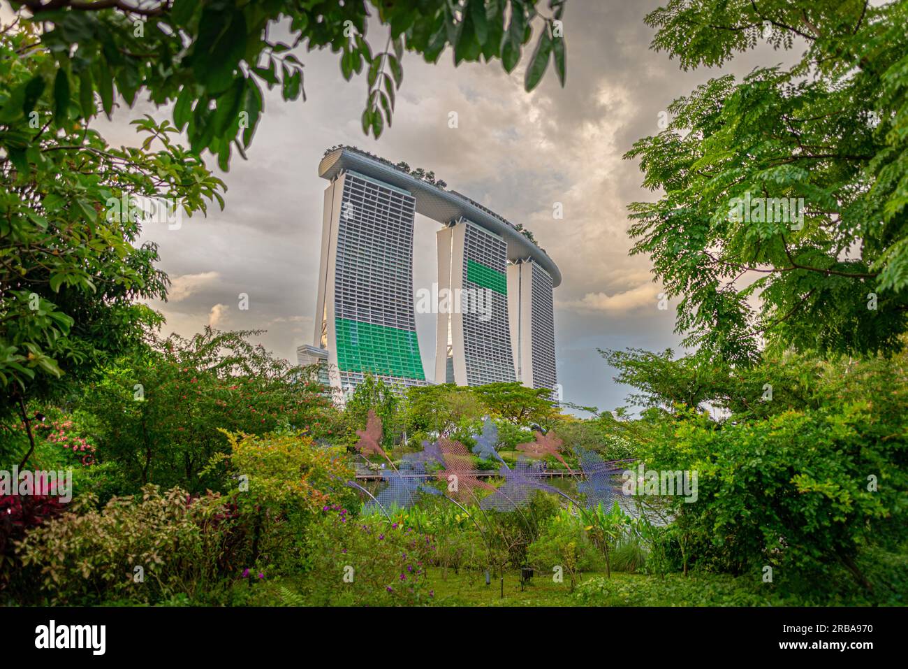 Una vista panoramica del complesso Marina Bay Sands incorniciata da vegetazione. Foto scattata dai vicini giardini del parco della baia in una giornata nuvolosa senza persone. Foto Stock