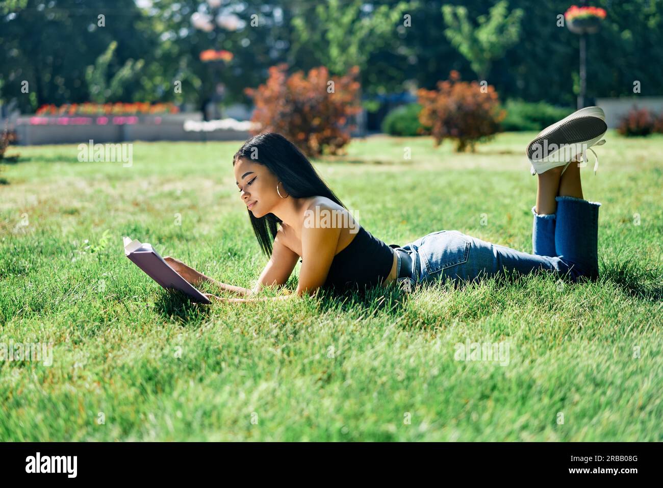 Bella donna afro americana leggere libro sdraiato in erba nel parco. Relax e hobby concetto Foto Stock