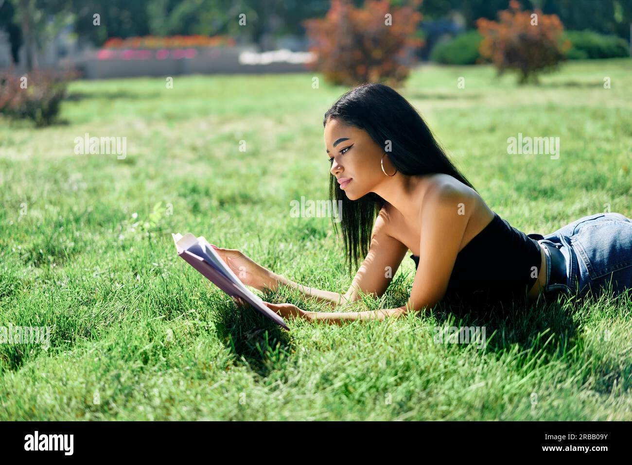 Piuttosto afro americano donna lettura libro sdraiato in erba nel parco. Relax e hobby concetto Foto Stock