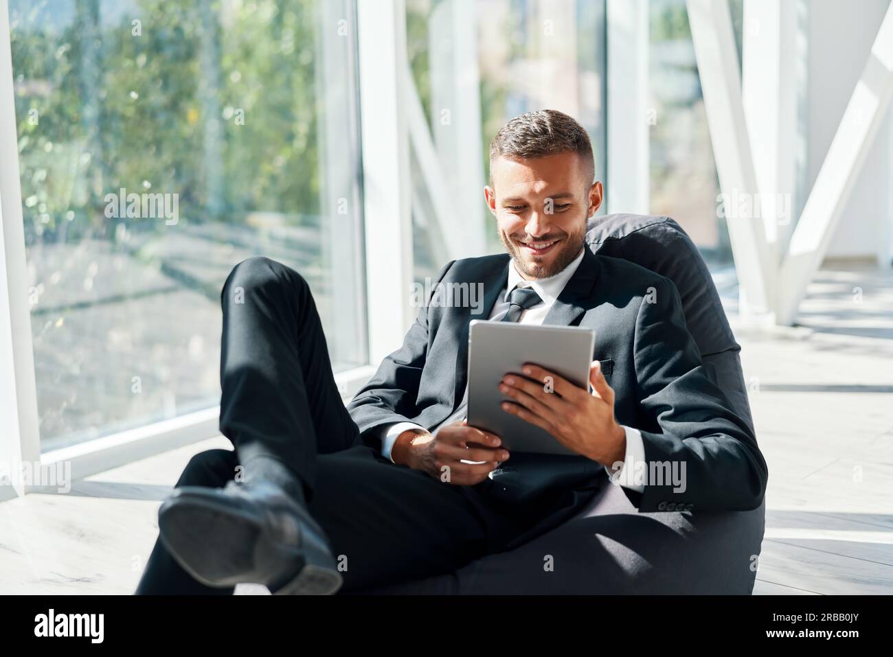 Sorridente uomo d'affari di successo in una suite elegante utilizzando un tablet seduto borsa chair fagiolo con finestre panoramiche di sfondo in moderno creativo ufficio Foto Stock