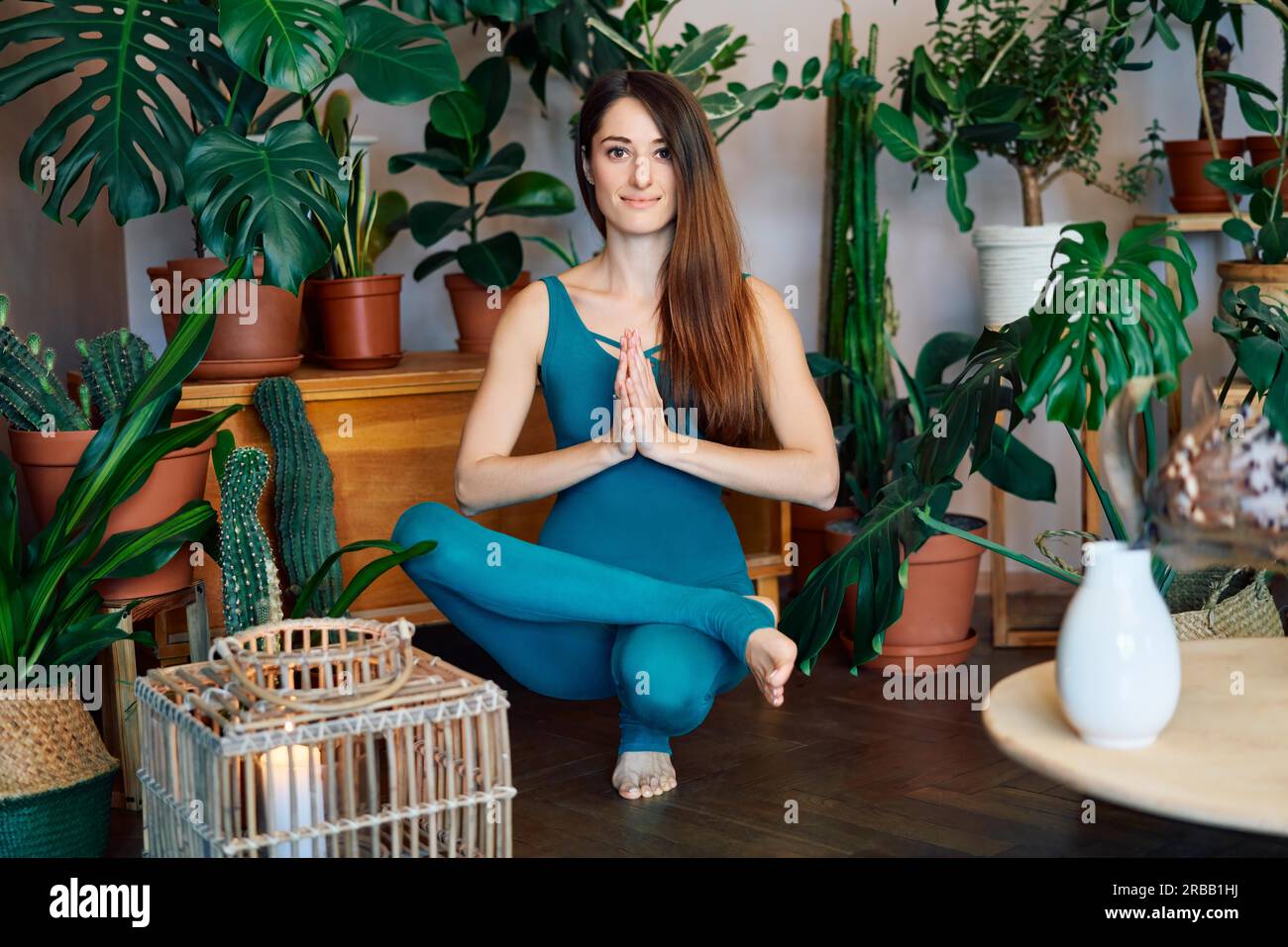 Giovane donna sportiva che pratica yoga facendo mezzo loto equilibrio piedi posa a casa. Padangustha Padma Utkatasana. Concetto di salute Foto Stock