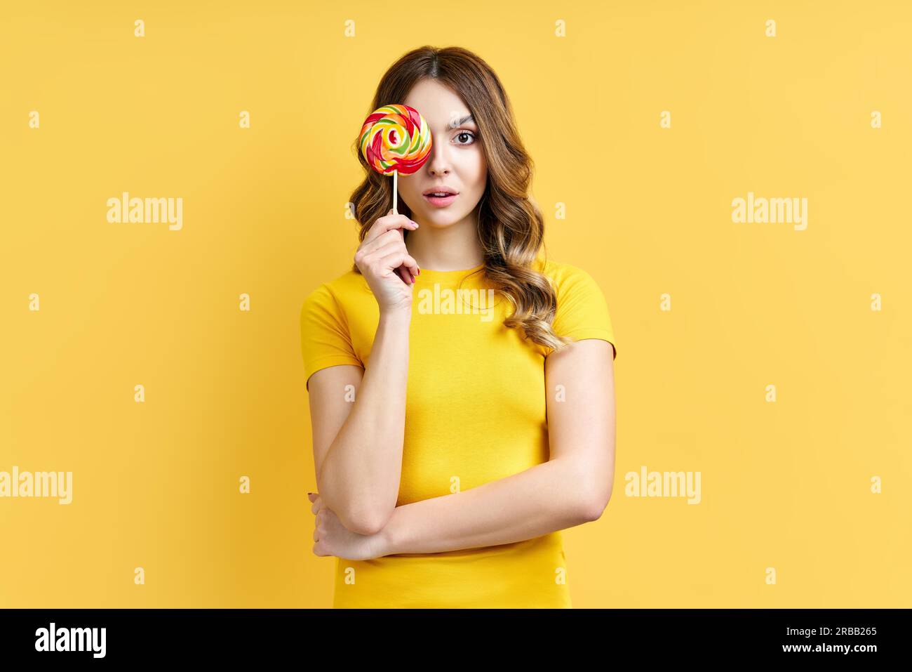 Bella donna copre il suo occhio con lollipop colorato isolato su sfondo giallo. Emozioni, divertimento, concetto di cibo Foto Stock
