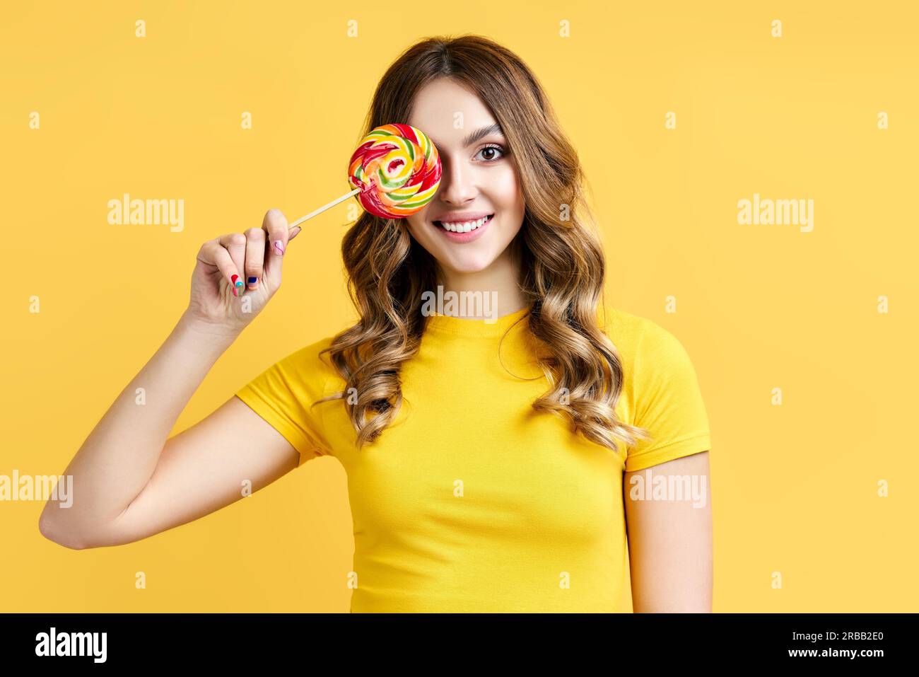 Bella donna sorridente copre il suo occhio con lollipop colorato isolato su sfondo giallo. Emozioni, divertimento, concetto di cibo Foto Stock