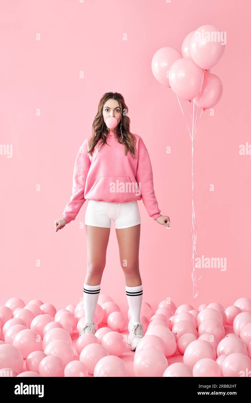 Giovane giocosa donna che soffia bolla con gomma da masticare che rimane in palloncini d'aria rosa sfondo. Festa, emozioni concetto Foto Stock