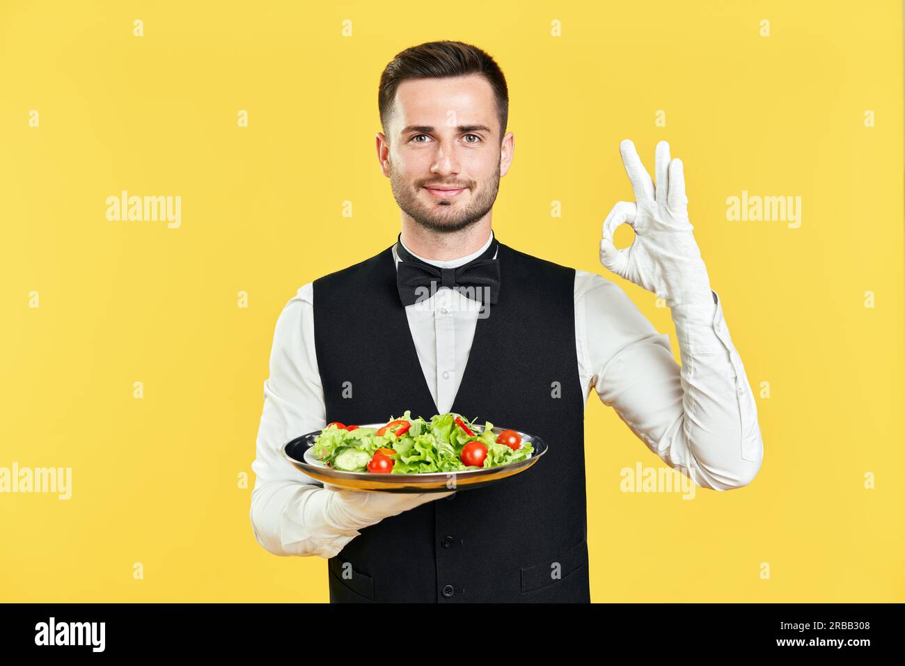 Sorridente sorridente piatto di tenuta con insalata di verdure salutari facendo segno ok con mano e dita su sfondo giallo. Dieta, ristorante, successo Foto Stock