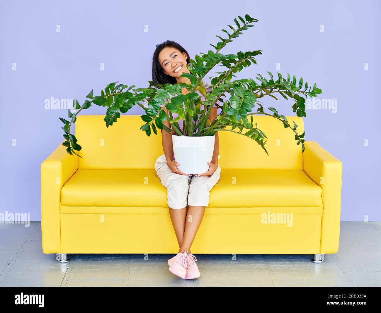 Giovane donna sorridente seduta sul divano che tiene enorme pianta in vaso. Divertimento, concetto di giardino domestico Foto Stock