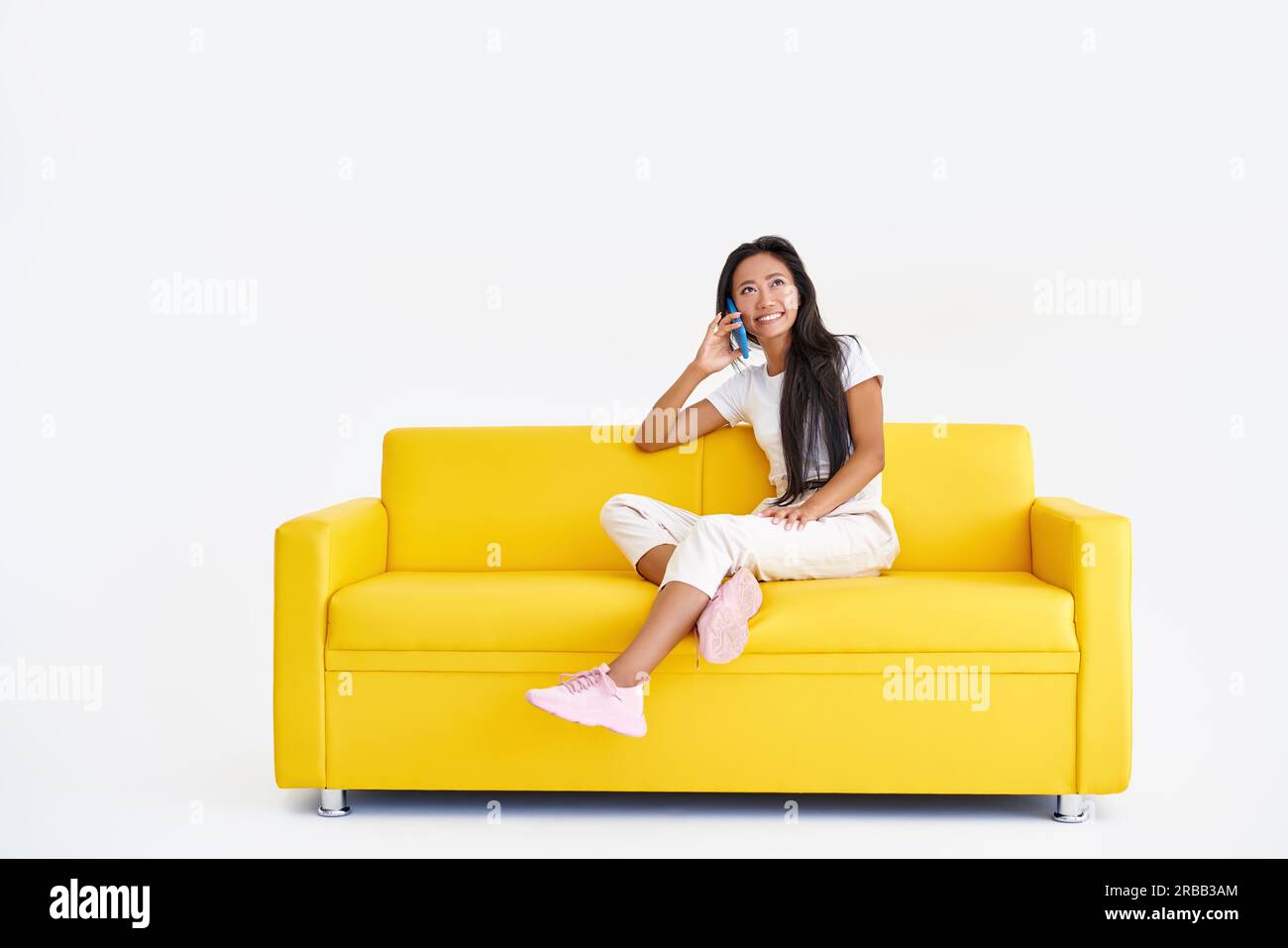Donna asiatica sorridente che parla al telefono cellulare seduto sul divano giallo su sfondo bianco con spazio copia. Tecnologia, concetto di comunicazione Foto Stock