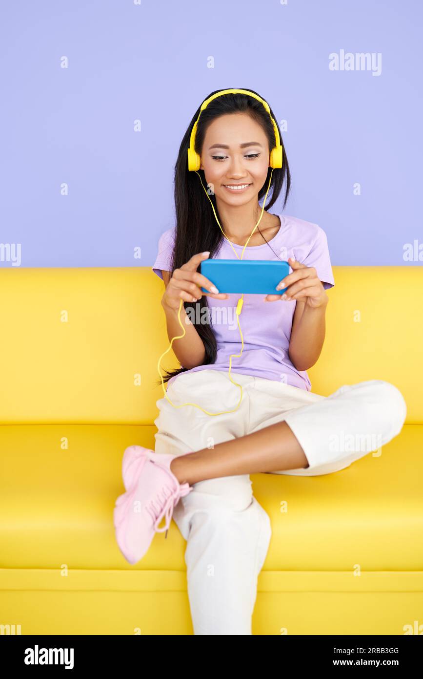 Donna asiatica sorridente che si gode le canzoni della playlist online in cuffia e naviga in Internet sul cellulare seduto sul divano a casa. Tecnologia, comunicazione Foto Stock