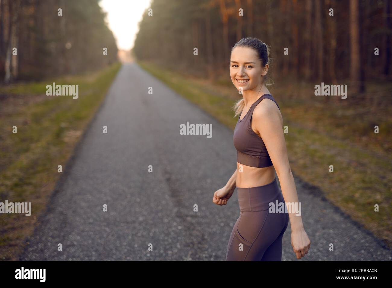 Allenati con una giovane donna atletica che fa jogging all'alba lungo una strada rettilinea che si allontana tra alberi boschivi con il bagliore del sole che alla fine si trasforma in un sorriso Foto Stock