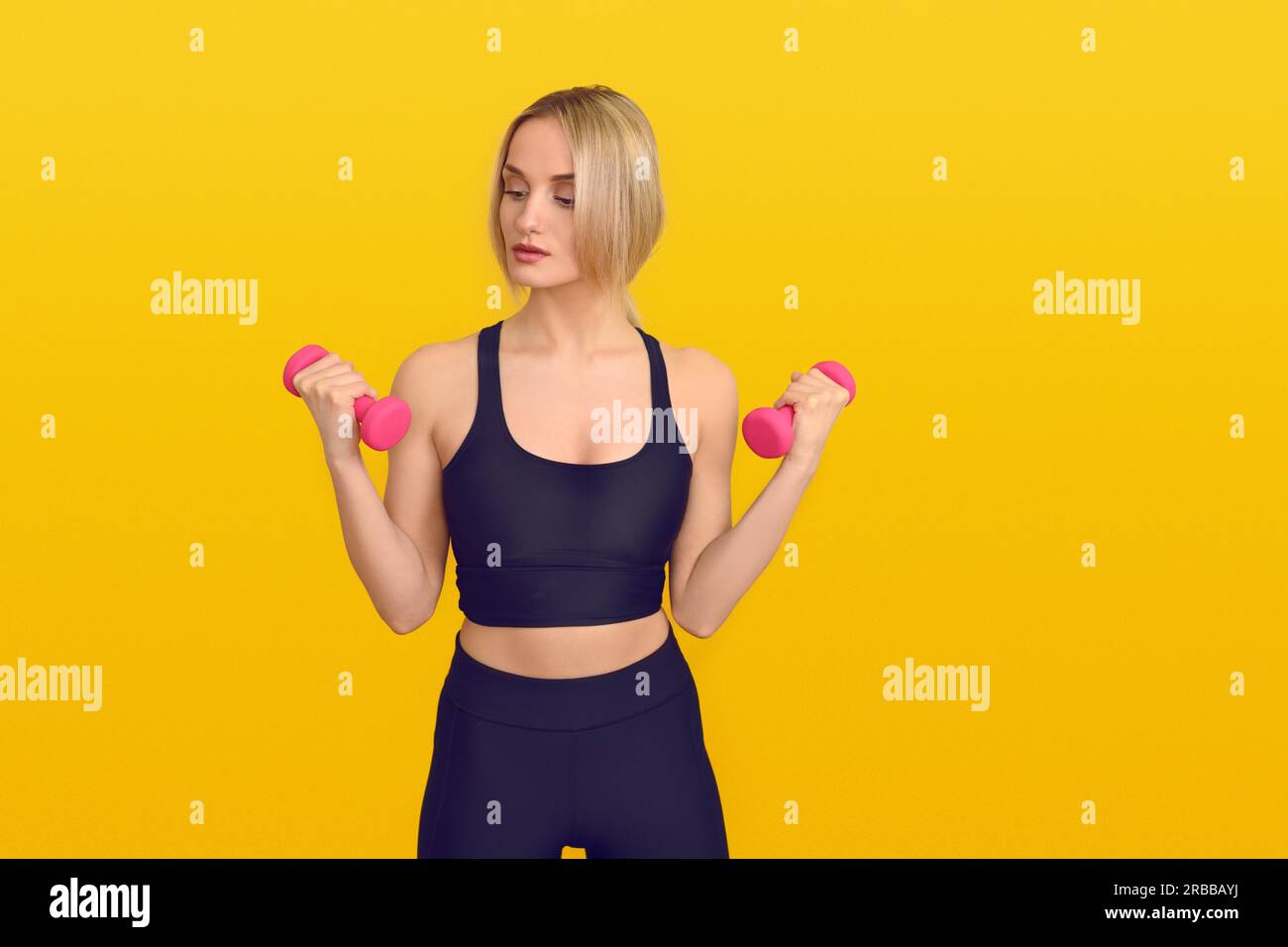 Giovane e bella donna bionda con abiti da allenamento neri in piedi con manubri rosa in mano su sfondo giallo chiaro con spazio per le copie Foto Stock