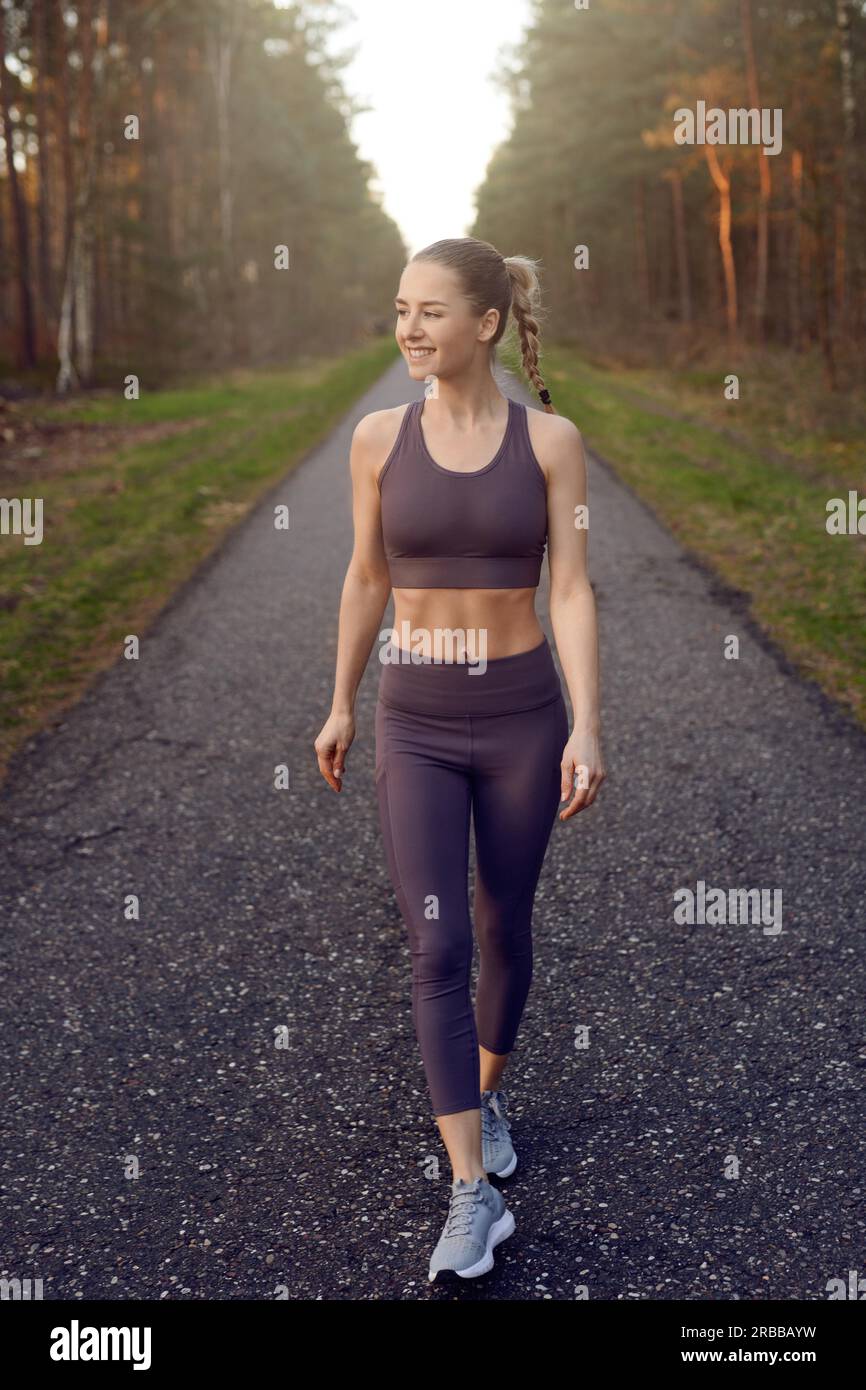 In forma giovane donna sana che cammina in abbigliamento sportivo su una pista forestale retroilluminata dal bagliore del sole al mattino o alla luce della sera in salute e fitness Foto Stock