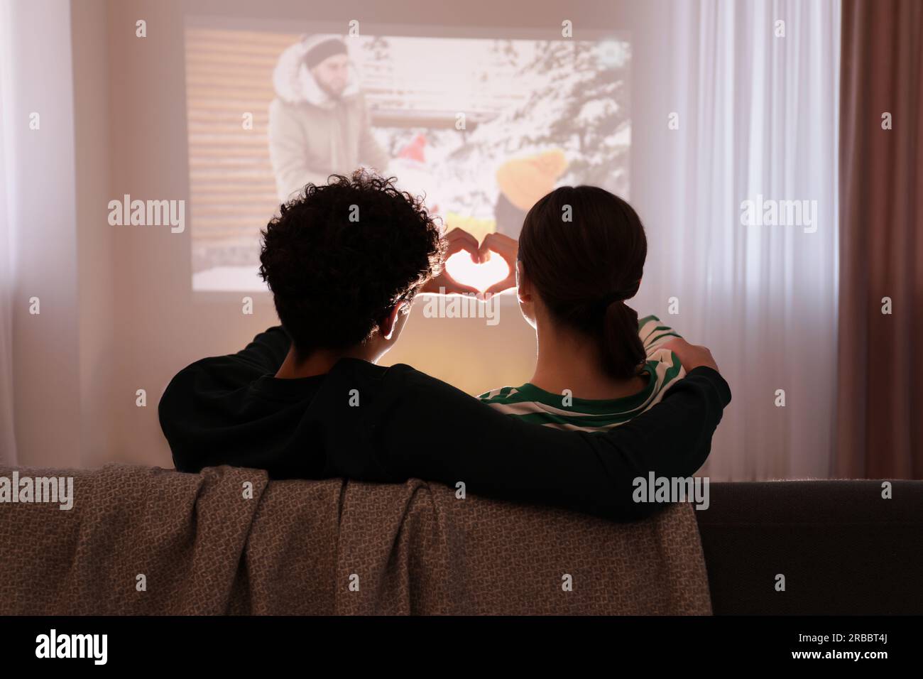 Coppia che guarda un romantico film di Natale tramite videoproiettore e che fa il cuore con le mani a casa, vista posteriore Foto Stock