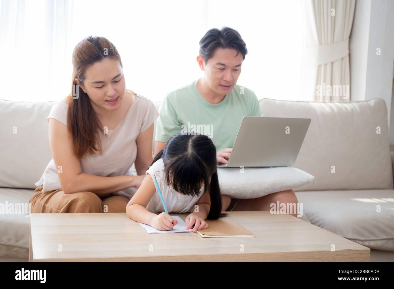 Famiglia felice con la madre che insegna i compiti a casa con la figlia e il padre che lavora con il portatile sul divano nel soggiorno di casa, mamma che spiega il lavoro scolastico con Foto Stock