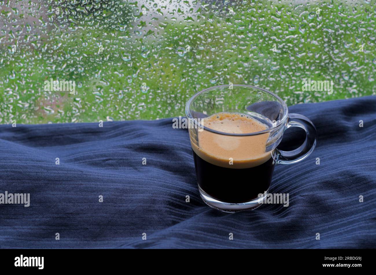 Un bicchiere di caffè espresso mette sul letto con goccia di pioggia sulla finestra durante la giornata piovosa. Concetto di "rimanere a casa". Foto Stock
