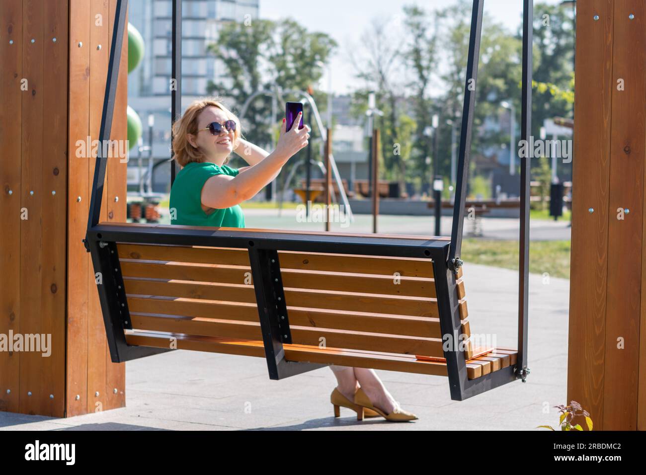 Una studentessa felice e attraente con gli occhiali da sole che scatta una foto selfie al telefono per strada. Una blogger seduta sulla panchina del parco e che usa lo smartphone Foto Stock
