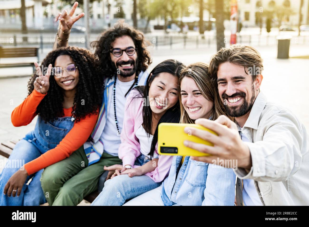 Diversi gruppi di amici che scattano un selfie in strada seduti su una panchina. Allegro gruppo multirazziale di giovani hipster che scattano una foto all'aperto in una Foto Stock