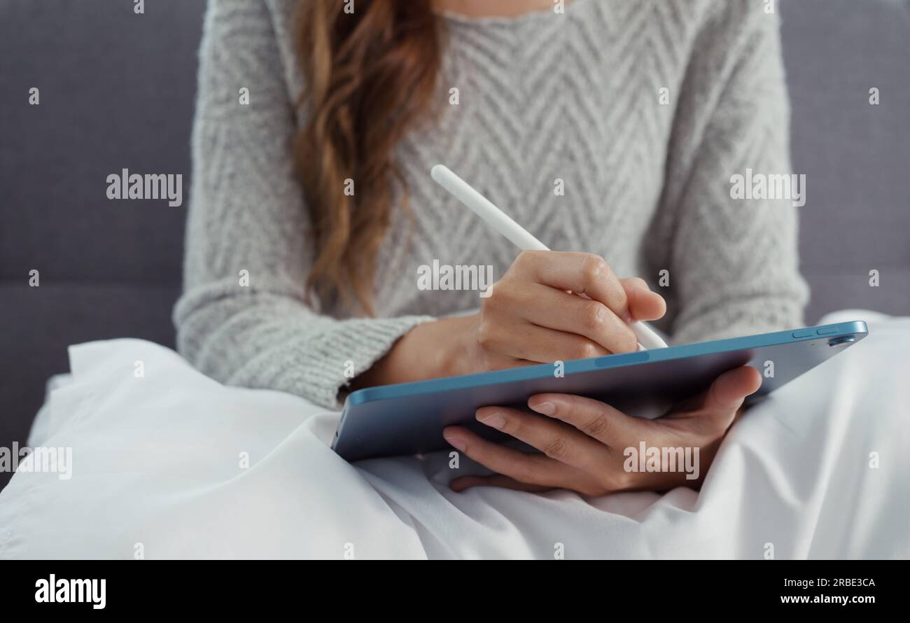 Giovane donna asiatica seduta su un divano che utilizza un tablet digitale e una penna per imparare a fare shopping o lavorare online a casa. Foto Stock