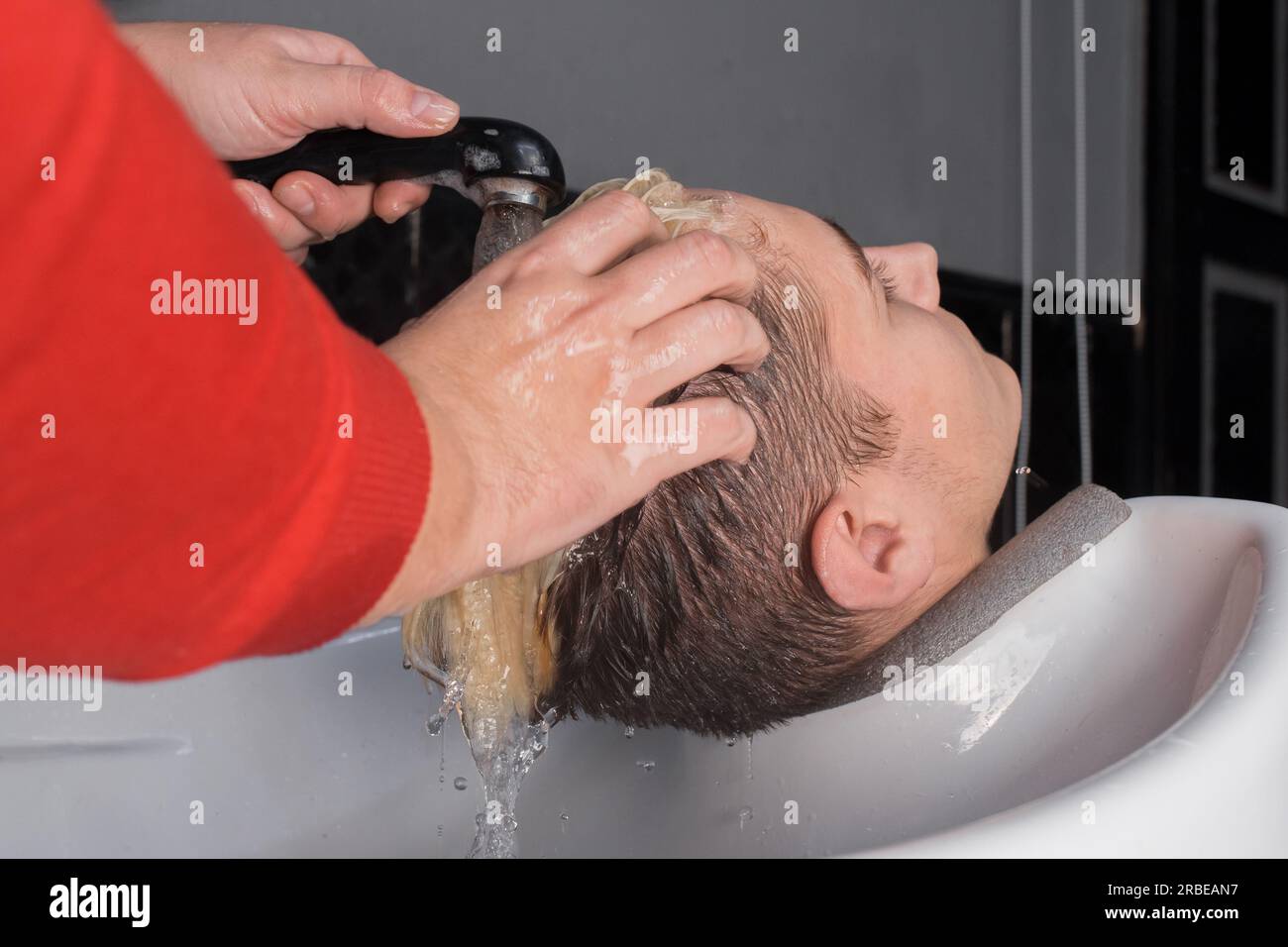 Le mani di un parrucchiere professionista si lavano i capelli con la pioggia con acqua a un cliente sopra il lavandino prima di tagliare i capelli. Foto Stock