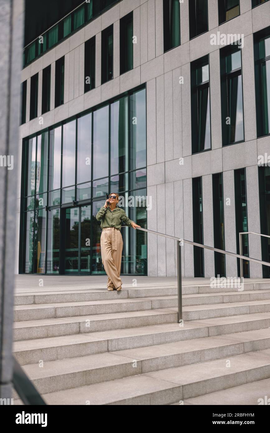 Una donna sorridente che lavora in ufficio parla al telefono mentre si trova sullo sfondo di un edificio moderno Foto Stock