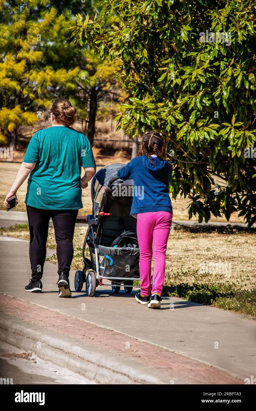 Madre bianca con figlia nera che spinge la carrozza del bambino che cammina su sentieri con alberi che circondano una messa a fuoco selettiva Foto Stock